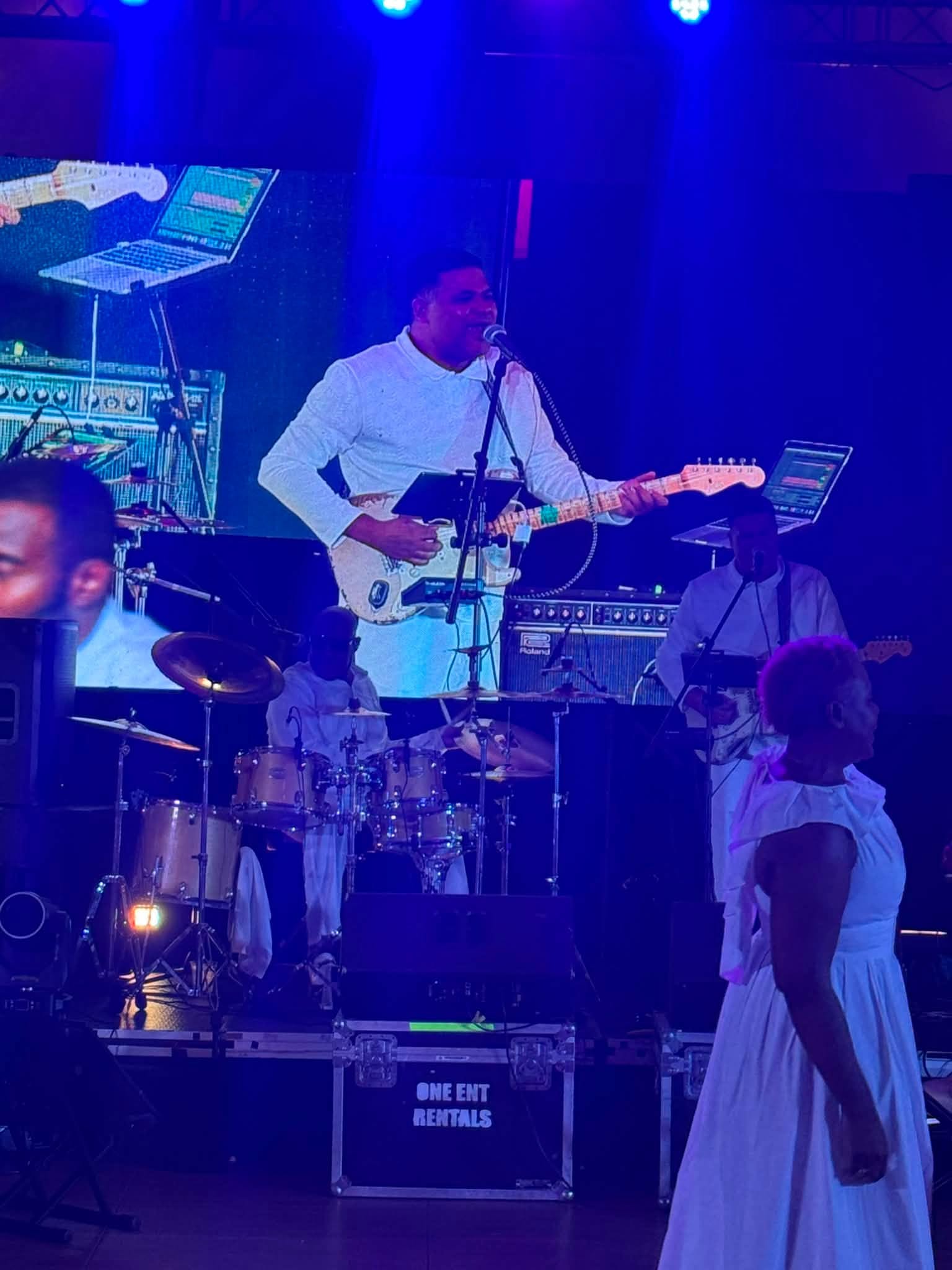 Ralph Conde1 - Caribbean Television Network Singer in white shirt playing a white electric guitar on stage, singing into a microphone under blue stage lights, with a drummer behind him.