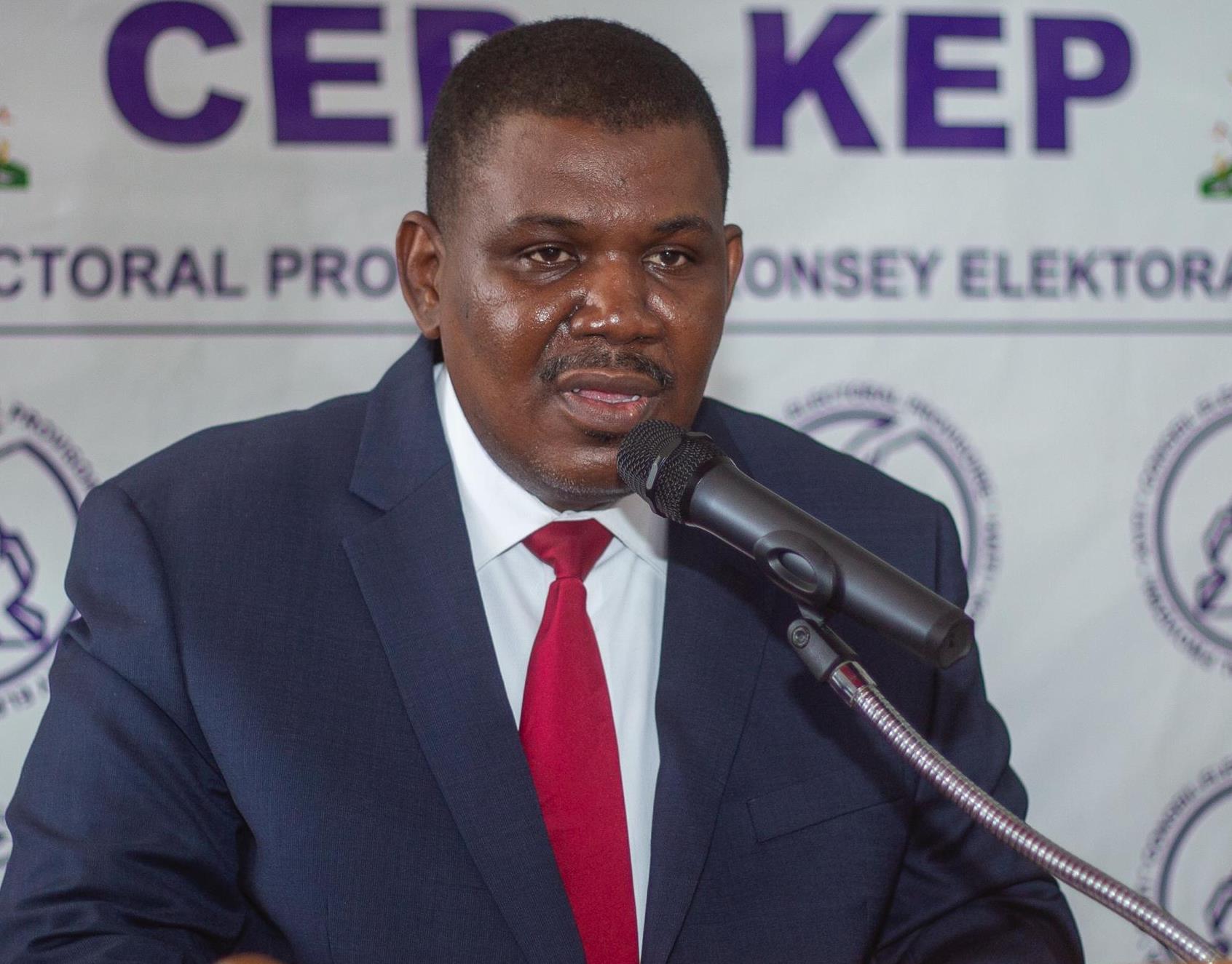 Man in a dark suit and red tie speaking into a microphone at a press event, backdrop with text and logos behind him.