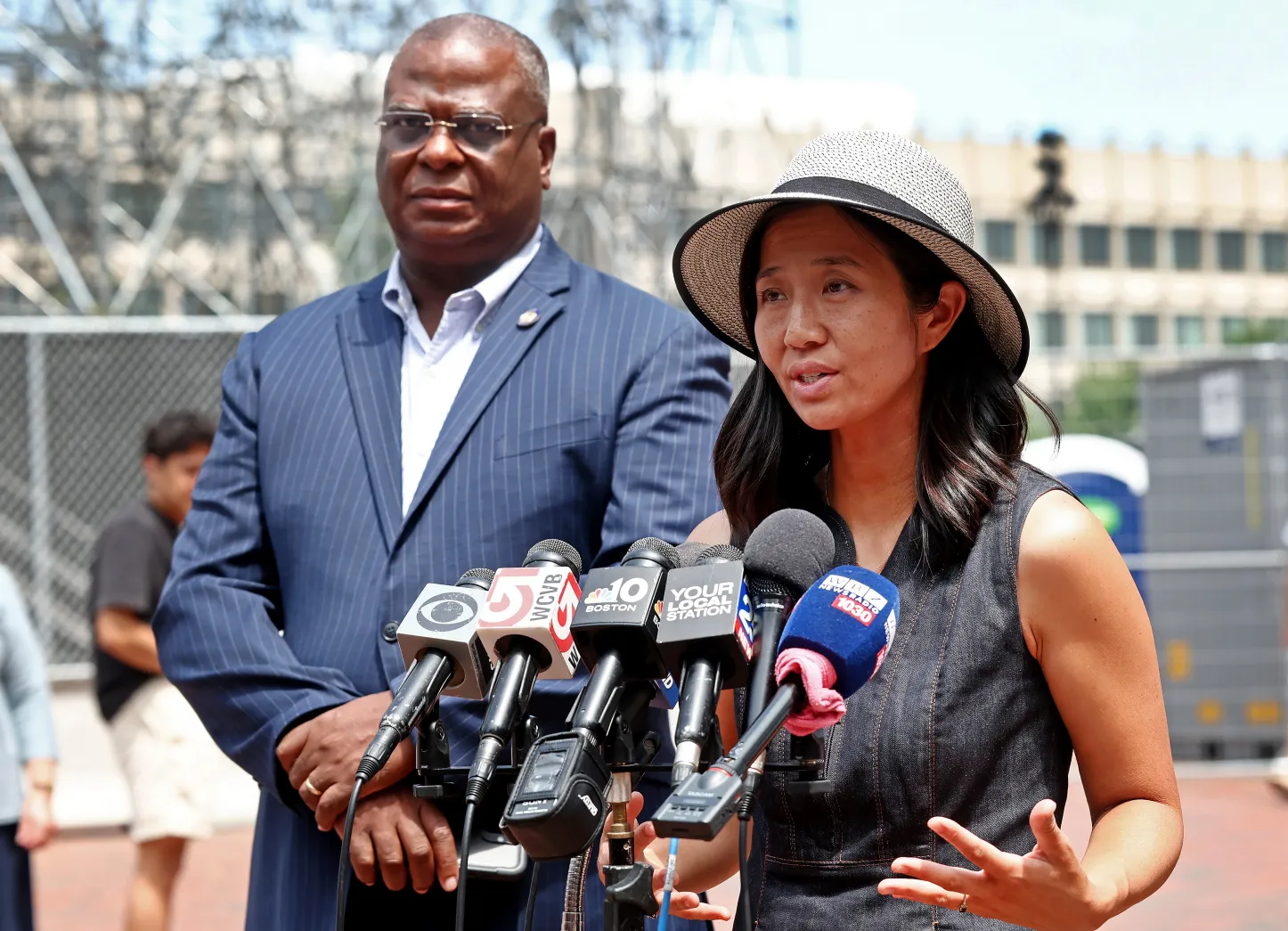 Woman in a hat speaks at a press conference with multiple microphones, while a suited man stands behind outdoors.