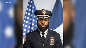 African American police officer in formal dress uniform, hat, standing with an American flag backdrop.