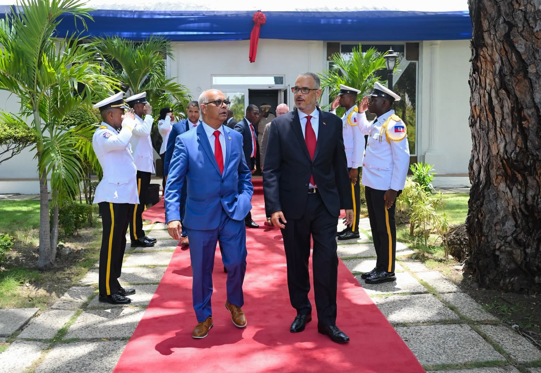 Two men in suits walk down a red carpet, flanked by saluting uniformed guards outside a white building with a blue awning.