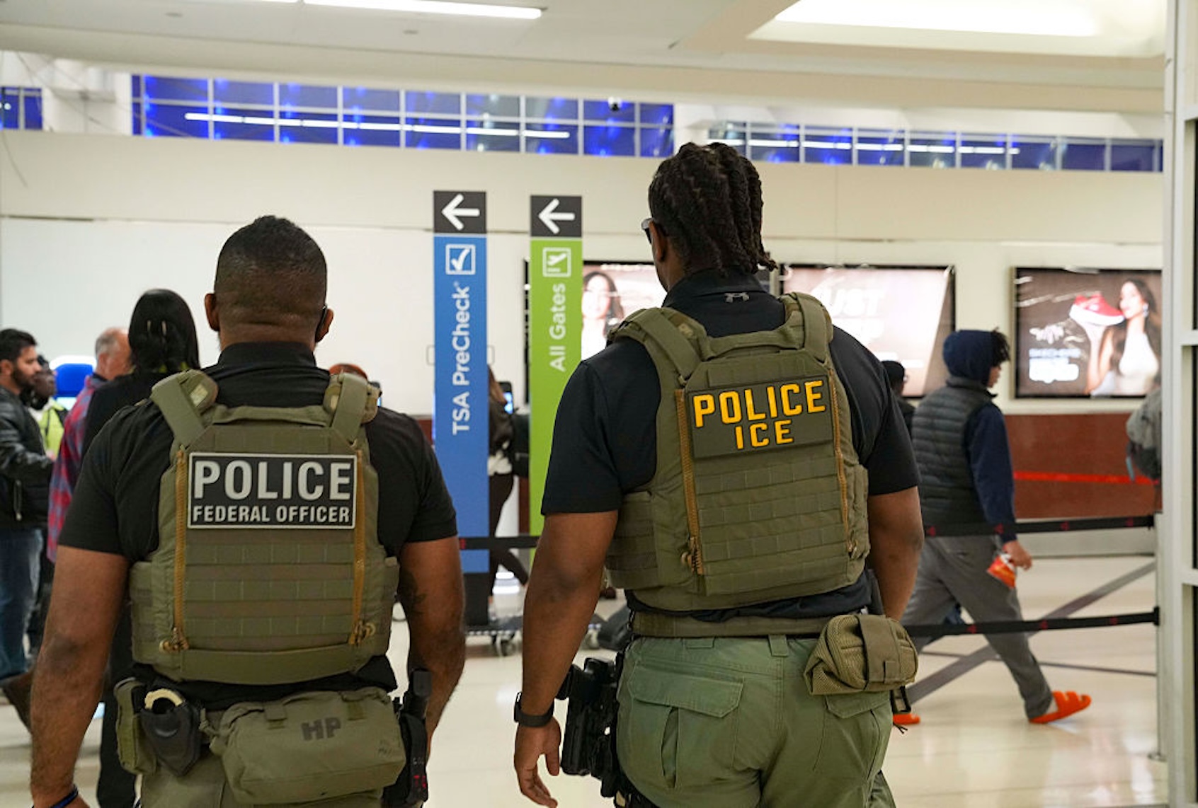 Two armed officers in tactical vests stand with their backs to the camera in an airport, POLICE and POLICE ICE visible on their gear.