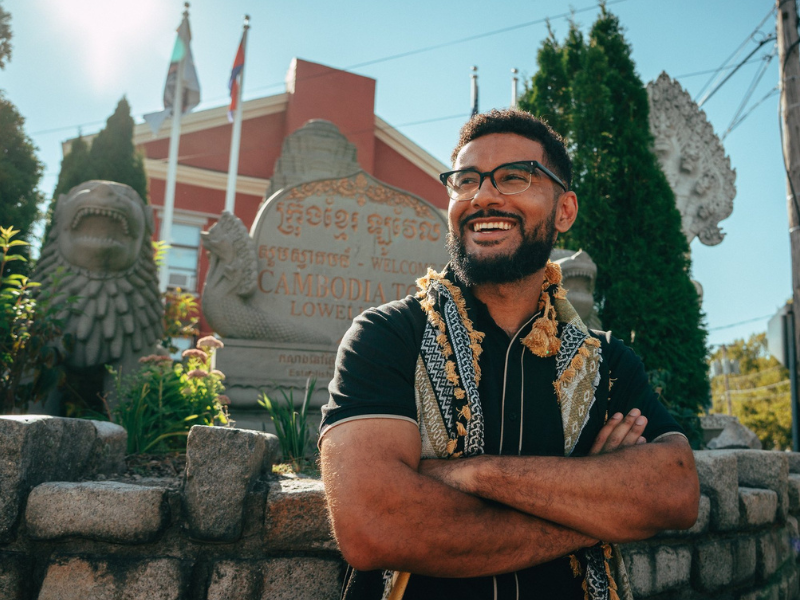 Smiling man with glasses and a patterned scarf stands with his arms crossed in front of a Cambodia welcome stone and stone lion statues.