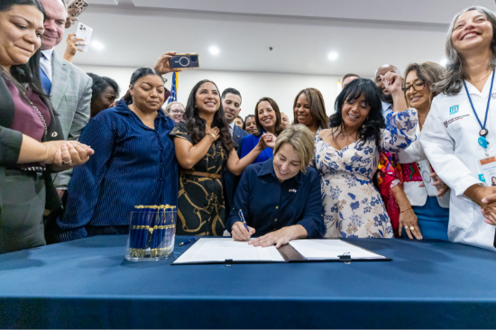 Woman seated at a blue table signs a document while a diverse group of colleagues watch and capture the moment on devices.