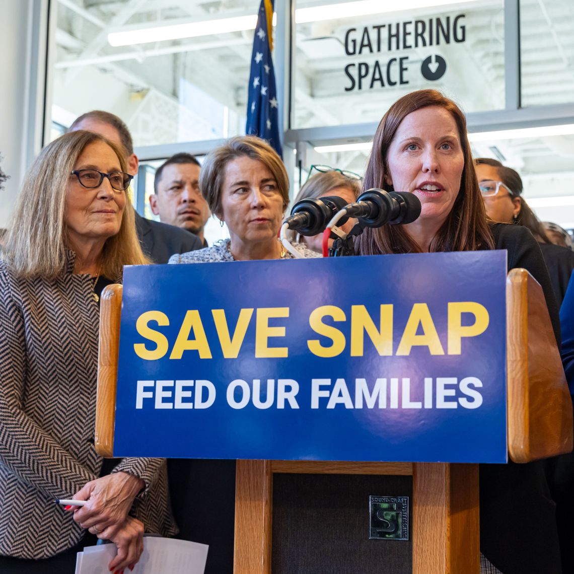 Woman at a podium speaking into multiple microphones, flanked by supporters; sign reads 'SAVE SNAP FEED OUR FAMILIES.'