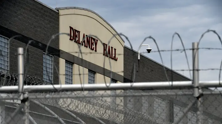 Exterior view of Delaney Hall behind a barbed wire-topped chain-link fence; a light-colored building with a curved façade and the Delaney Hall sign visible.