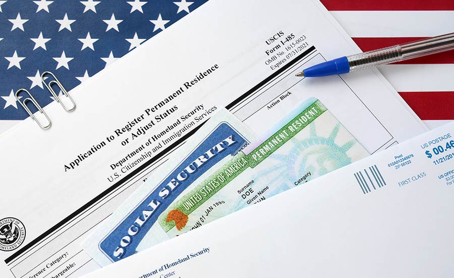 Immigration paperwork with a US flag background and a green permanent resident card (green card) on top of forms with a pen nearby